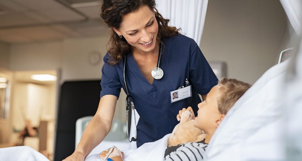 Nurse in blue scrubs smiling at a young boy holding a teddy bear in a hospital bed