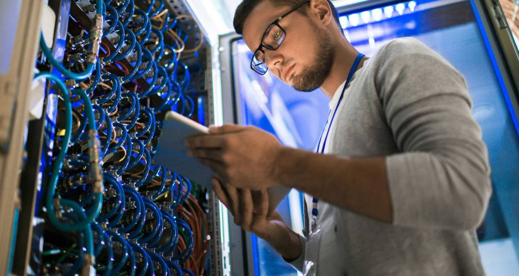 Man in glasses checking a tablet beside cabled server racks in a data centre