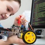 Girl testing a small robot car's circuit board with a probe beside a screen of code