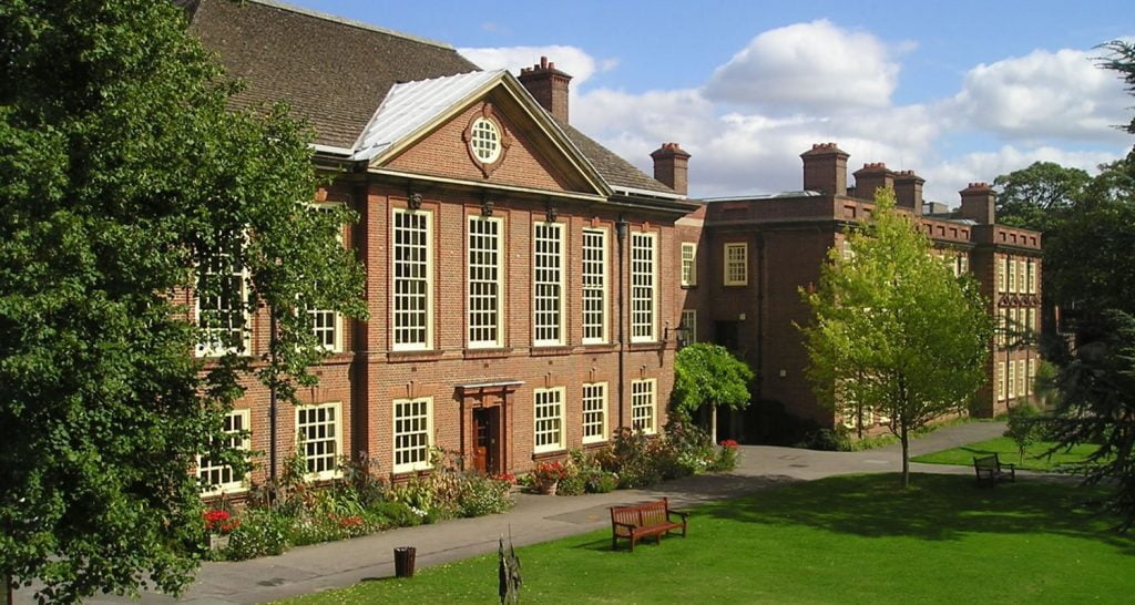 Large red-brick building with tall windows beside a sunny lawn and benches