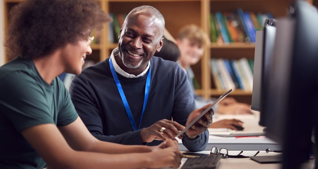 Smiling teacher showing a student a tablet at a computer desk in a library
