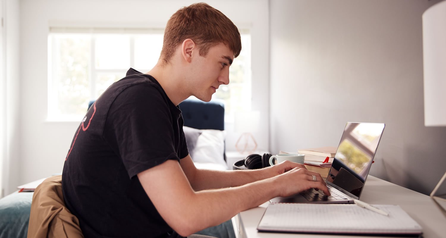 Student typing on a laptop at a desk in his bedroom