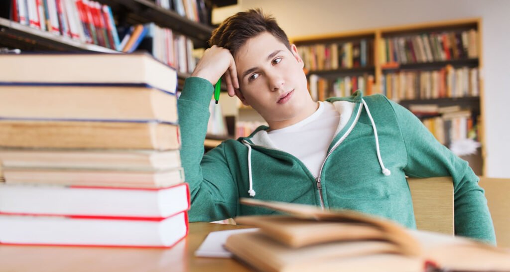 Tired student leaning his head on his hand beside a tall stack of books in a library