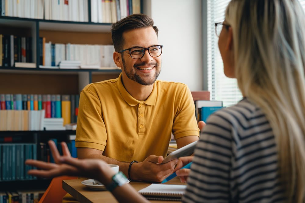 Smiling man in glasses talking with a woman across a table in a room lined with bookshelves
