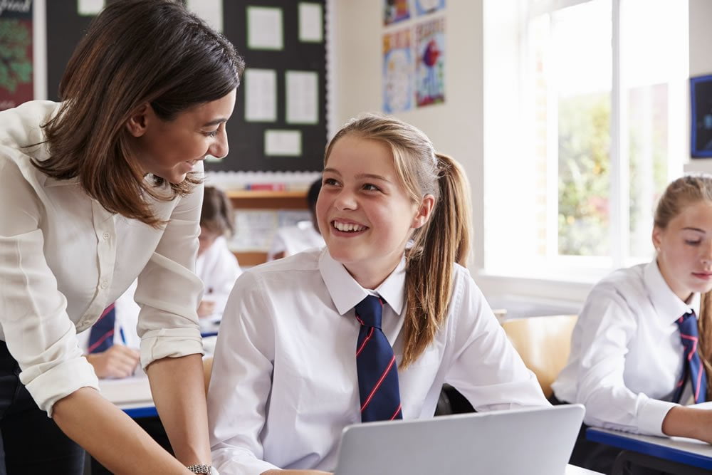 Teacher smiling with a pupil who is working on a laptop in a classroom